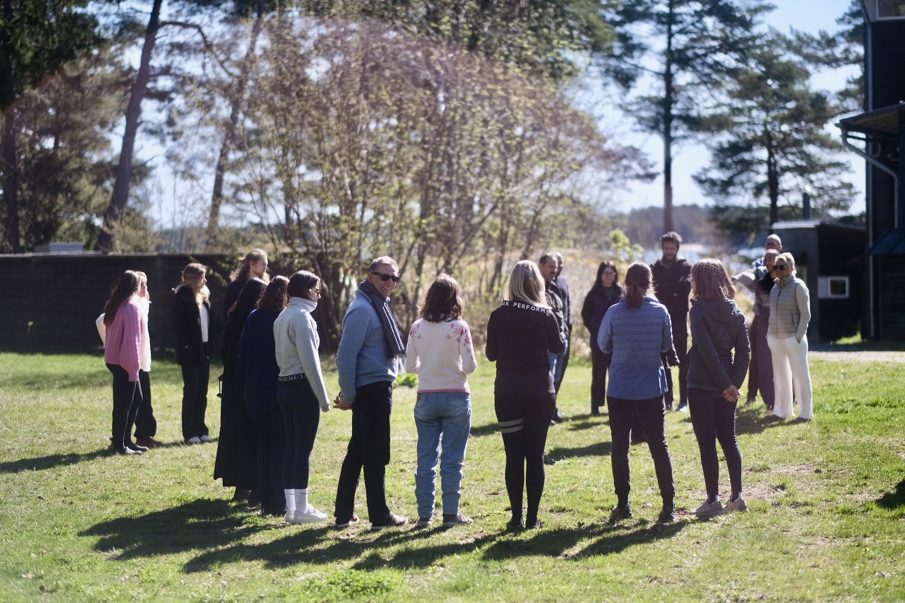 Participants standing in a wide circle on the grass between the pines